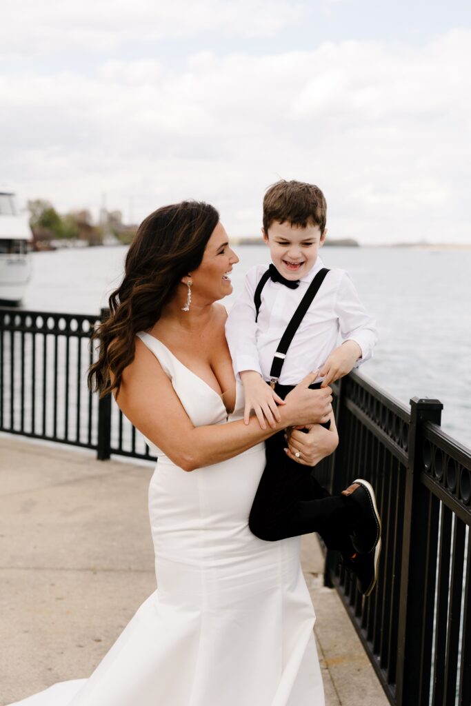 the bride smiles while holding her son on a dock in front of the water